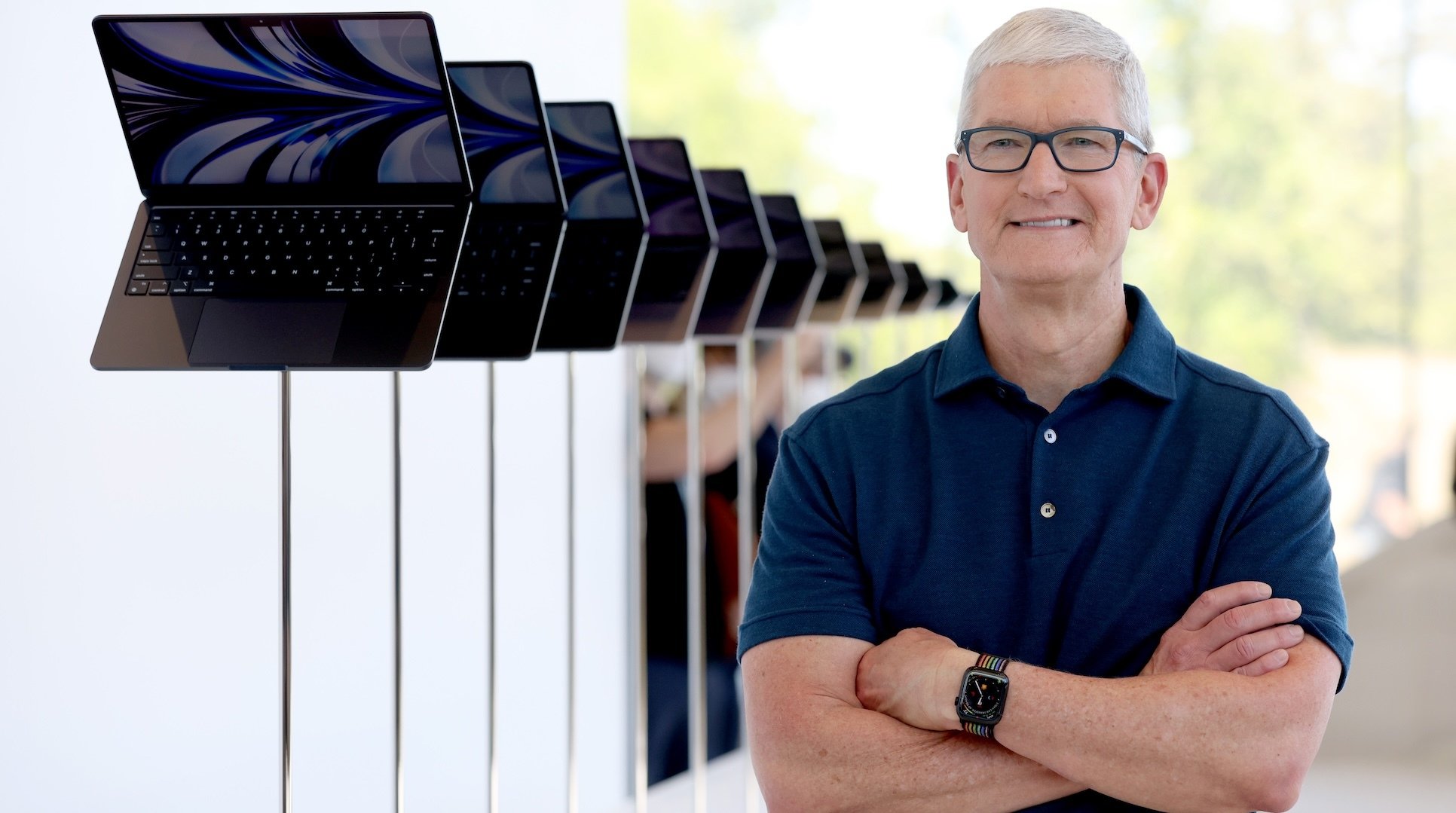 Tim Cook smiling, arms folded, next to a display of Macbook Airs