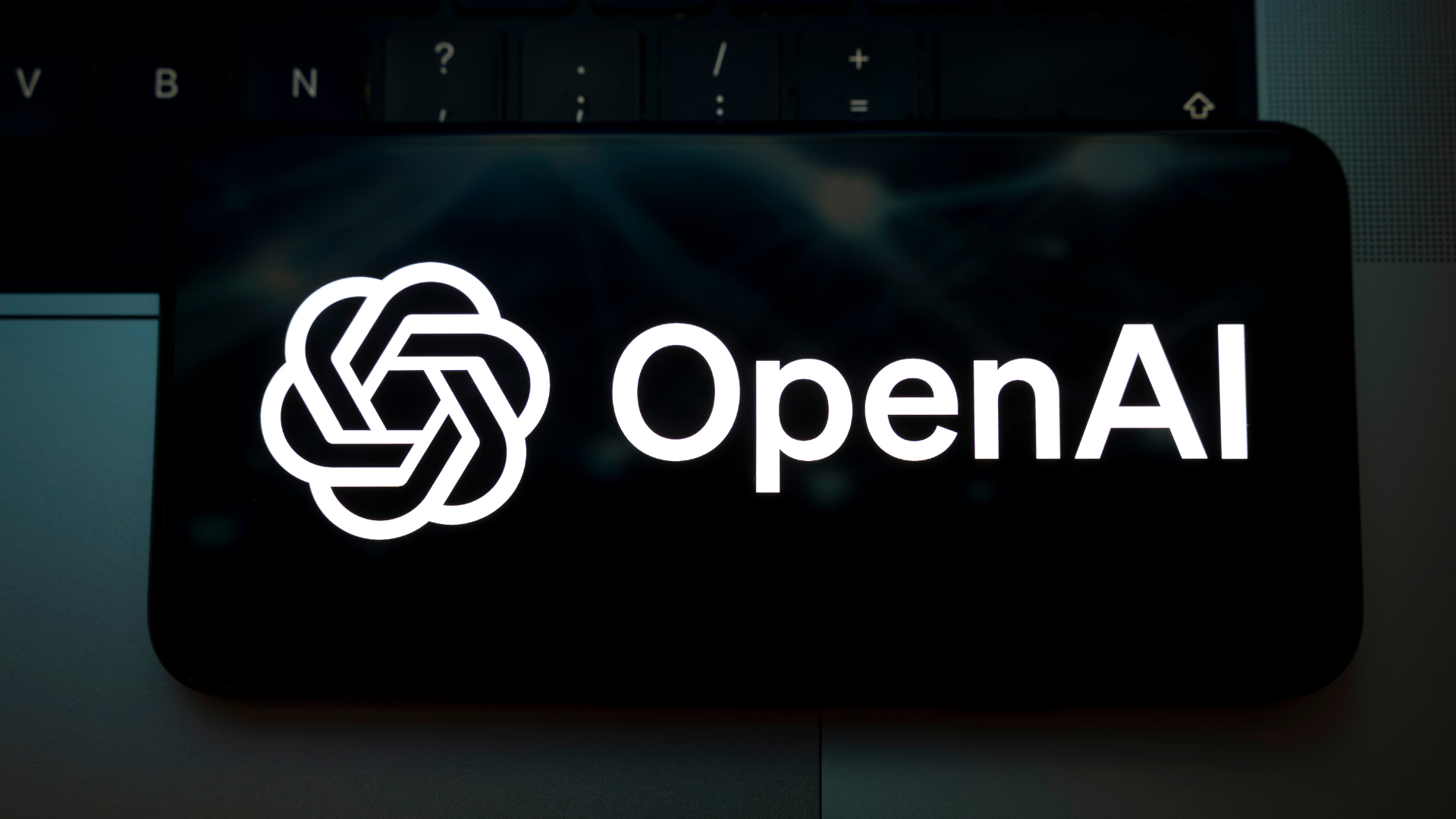 The OpenAI logo appears on the screen of a smartphone placed on a laptop keyboard illuminated by green light.