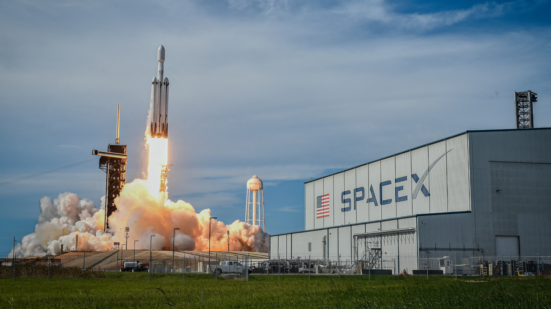 A SpaceX Falcon Heavy rocket lifting off, next to a building bearing the SpaceX logo.