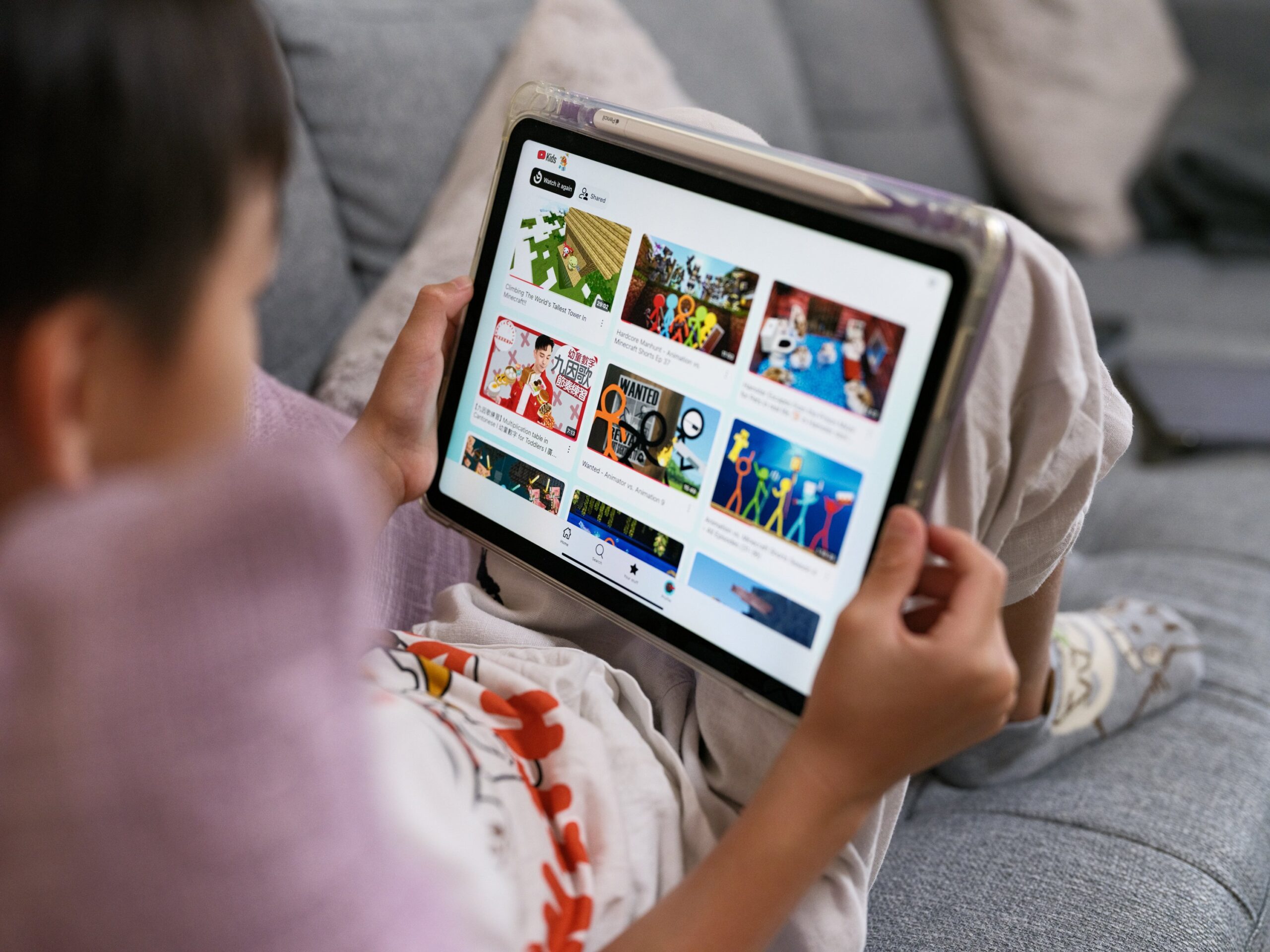 A child sits with an iPad resting on their knees. The screen shows the YouTube Kids homepage.