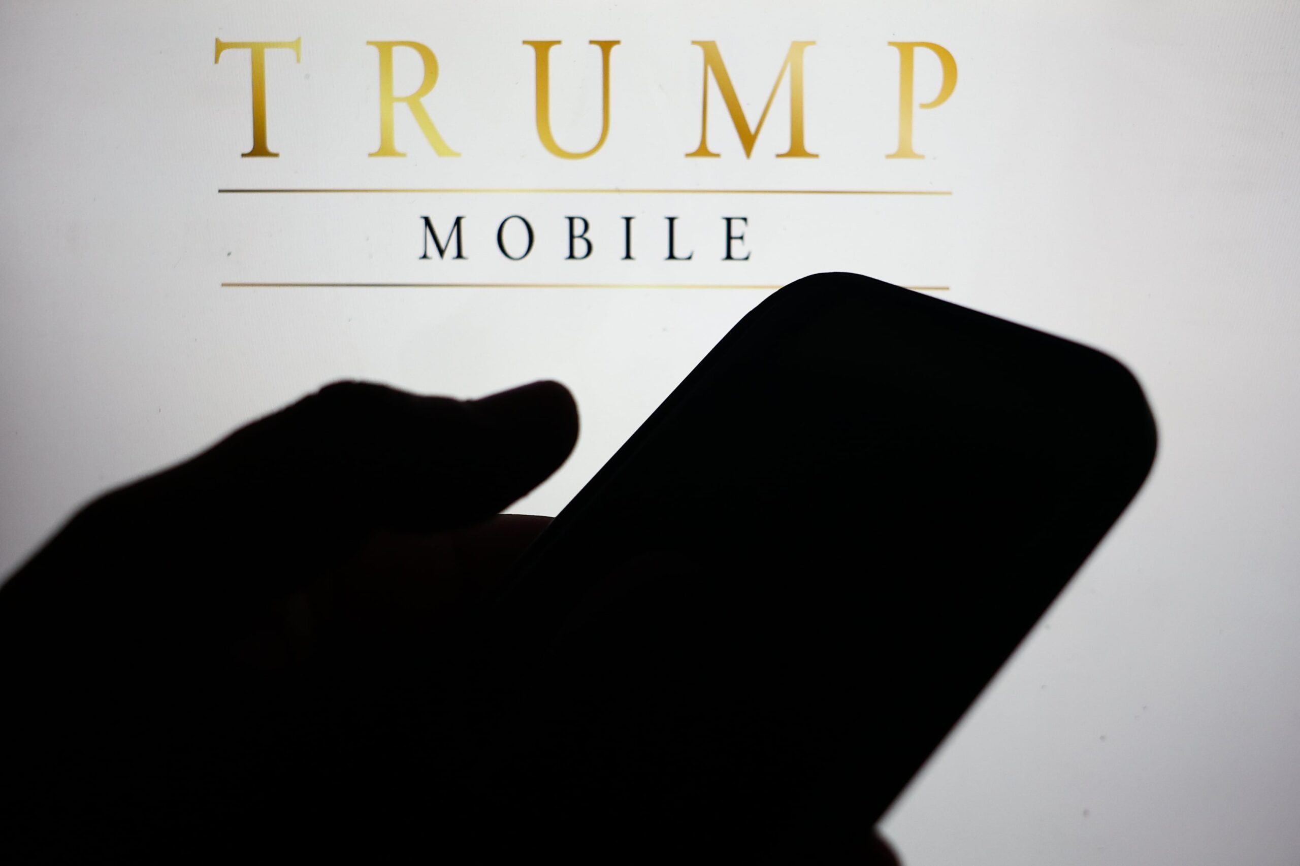 A shadow of a hand holding a phone in front of the Trump Mobile logo.