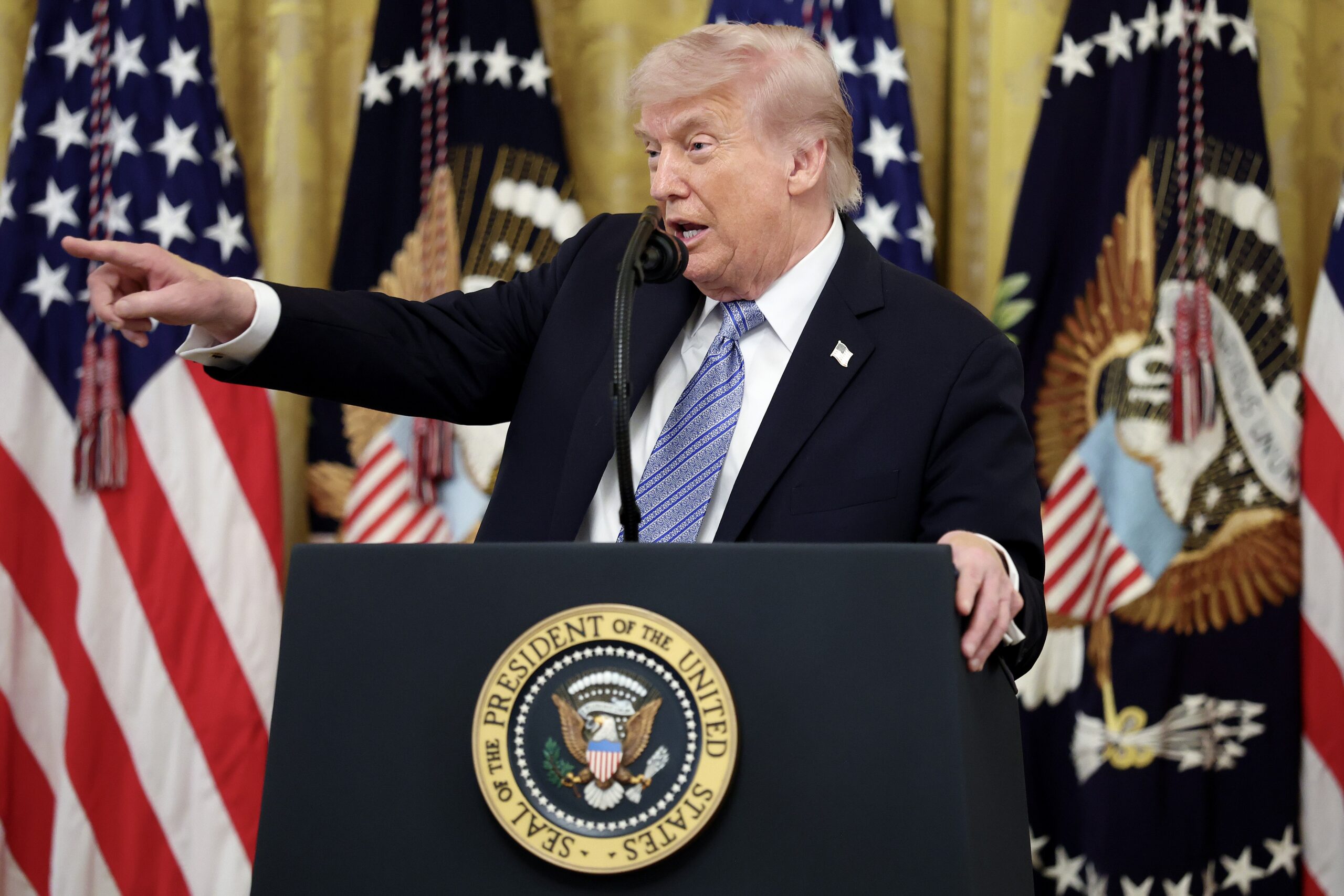 Trump stands at a White House podium pointing to the right. 