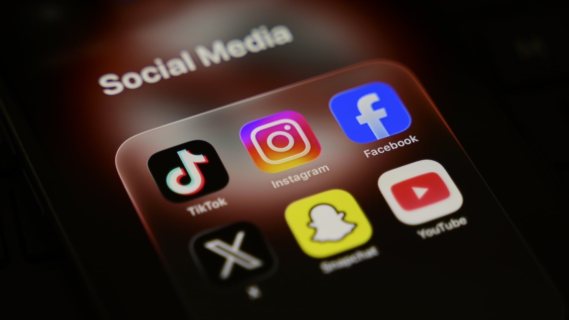A smartphone displays a folder of social media applications, including TikTok, Instagram, Facebook, X, Snapchat, and YouTube.
