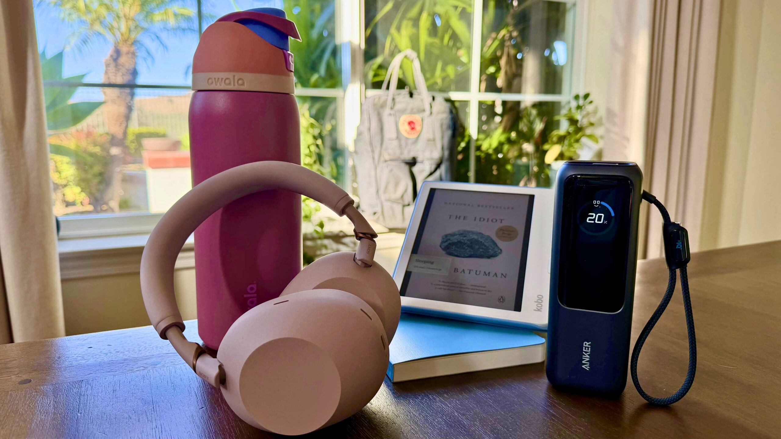 headphones balanced on water bottle next to portable charger, notebook, and kobo on the edge of a table