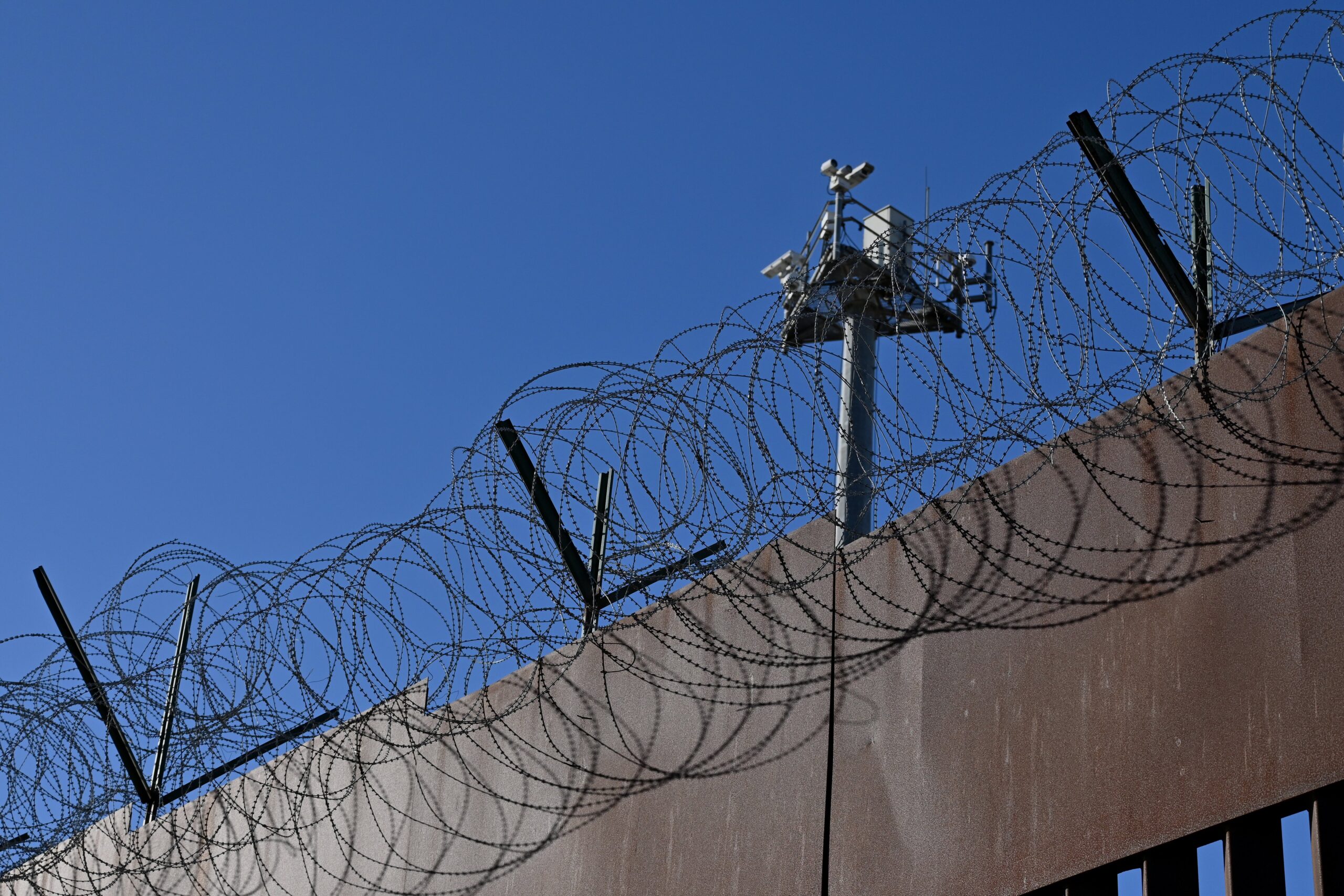 A surveillance tower looms behind a concrete wall with barbed wire on top. 
