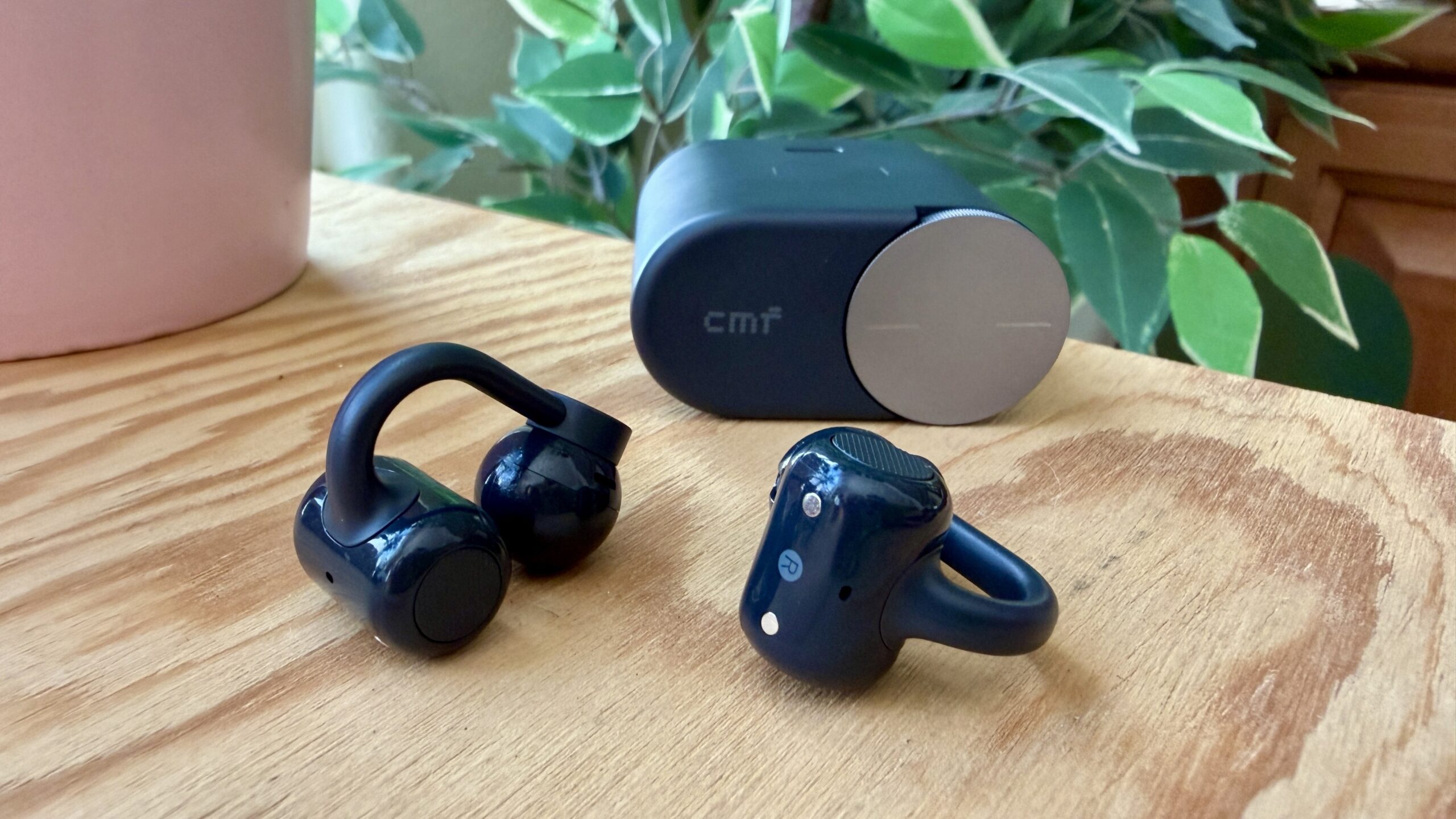 dark grey cmf clip pro earbuds on wooden tabletop in front of pink vase and a houseplant
