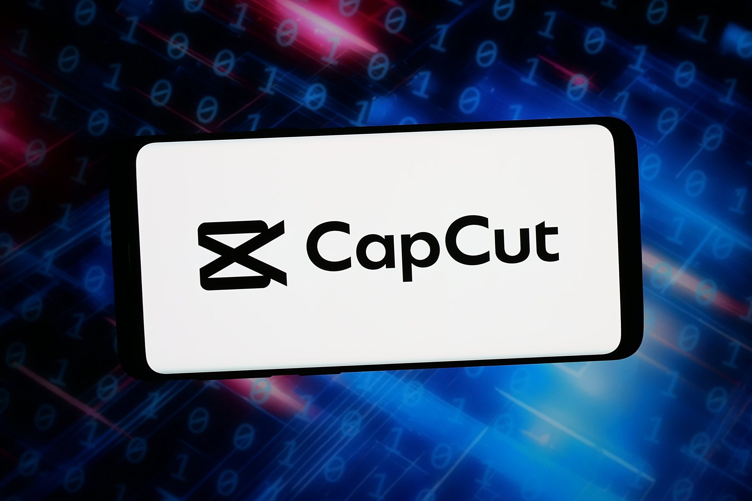 , the logo of video editing app CapCut (JianYing) is seen displayed on a smartphone in front of an abstract background on computer screen
